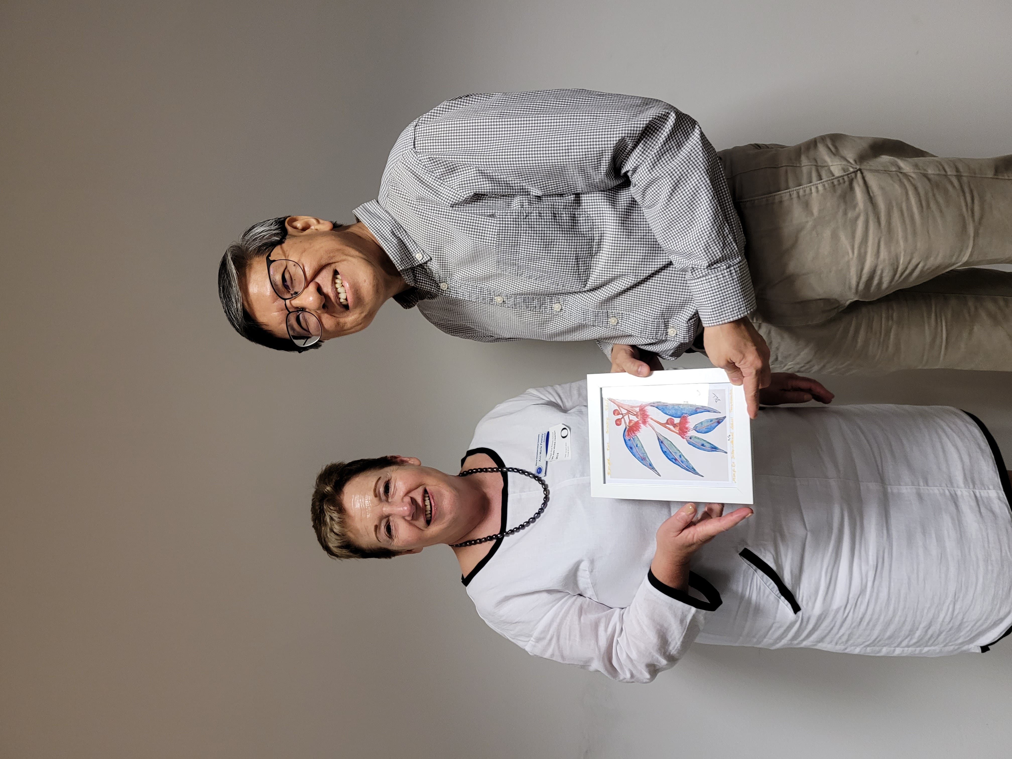 Two smiling people stand together holding a small framed watercolor painting of a floral branch with a red figure.
